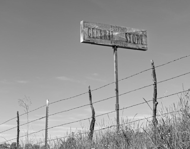 A black and white landscape photo that is a view looking up toward a barbwire fence at the top of a hill. A bit of long grass is seen in the lower right. Then the fence and a clear blue sky in the background. A very old sign is on the opposite side of the fence on the right. It is a wood rectangle about three feet long by one foot wide. It is mostly gray from weathering but there is evidence of white paint. In very faded black lettering it says, "Hogland, General Store, Est 1920."