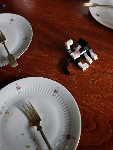 A photograph showing a wooden table top with three used plates with small forks next to a small black-and-white LEGO-style cat cleaning itself.