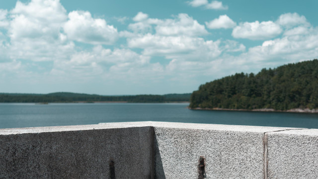 The corner of a granite block wall is in the lower foreground. Above that a reservoir with a headland in the mid-ground camera right, and the far shore at the horizon. The sky is partly overcast with fair weather clouds. The sun is nearly overhead, casting hard shadws on the granite wall, and revealing its texture.