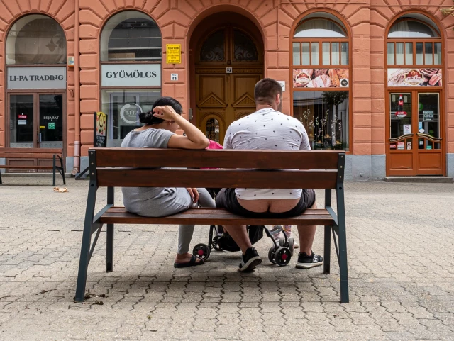 Fußgängerzone, Parkbank mit Männlein und Weiblein, davor ein Kinderwagen. Bank in Menschen mit dem Rücken zum Betrachter.
Männlein zeigt überzeugend sein Bauarbeiter Dekolleté.
Gegenüber ein Gebäude mit Obst- und Gemüseladen.