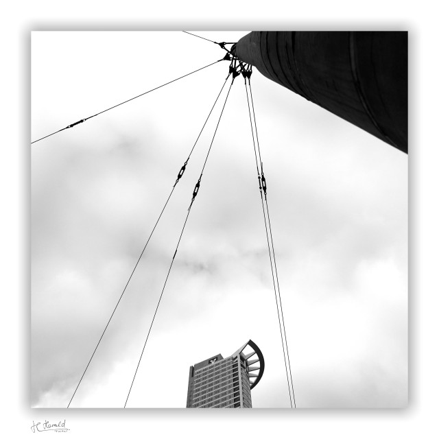 Square black-and-white image of a cloudy sky with only a few cloud formations visible. In the upper right corner, the top part of a large steel post protrudes into the image, to which six wire ropes with tensioning devices are attached, running to the left and down out of the image. The top of the West End Tower protrudes into the image from below.