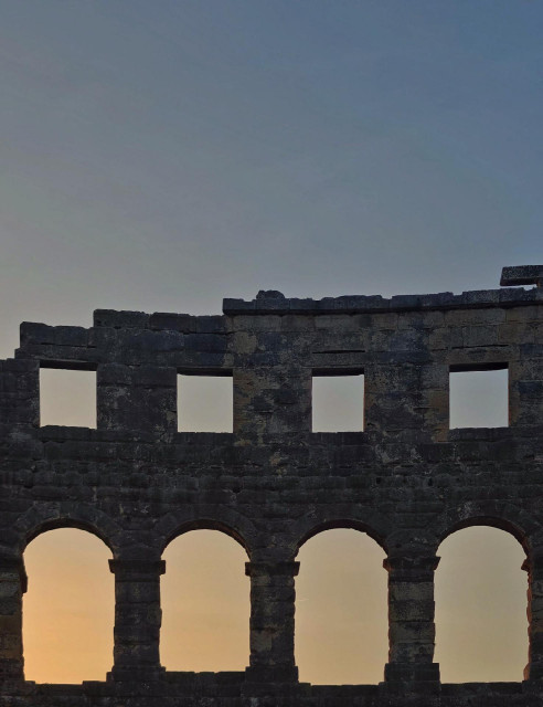 An evening photo of an old stone wall with rectangular and arched openings that belongs to a ruin of an amphitheater. A sunset sky with a soft gradient can be seen through the openings in the wall.