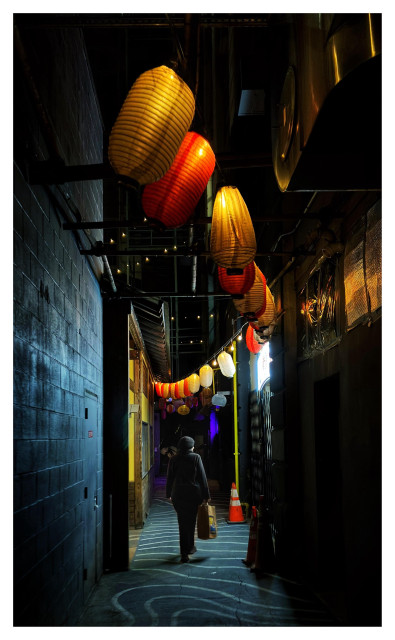 Nighttime photo of a narrow alley illuminated by hanging Japanese lanterns in alternating colors of red and yellow. The gray pavement is marked with wavy white stripes. A person in a dark winter hat and coat stands with their back to the camera in the middle distance, carrying a paper shopping bag and illuminated by the light from a restaurant storefront on the right side of the alley.