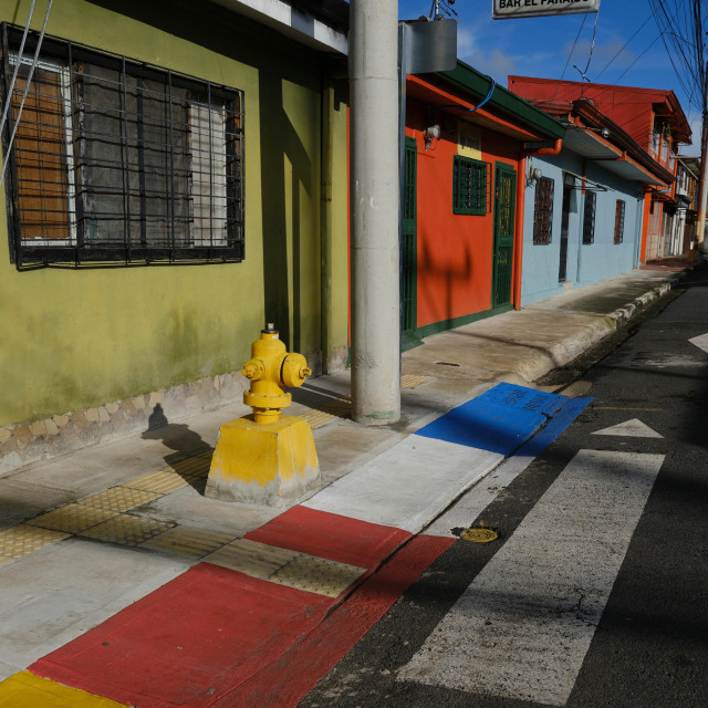 Square color photograph of street corner in Costa Rica. Perspective lines interact  with painted color bands and projected shadows, to create a complex structure and visual plane.