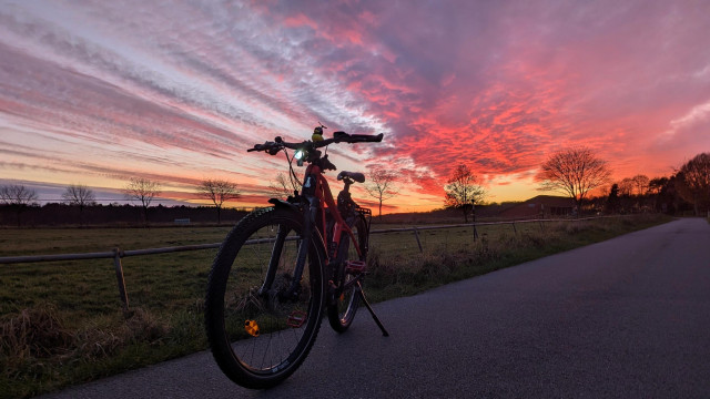 Ein rotes Raleigh Dundee MTB steht auf einem Wirtschaftsweg nach Sonnenuntergang. 
Der Himmel ist orange, lila, pink, rot, blau. 
Im Hintergrund stehen einige kahle Bäume.