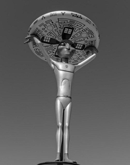 A black and white portrait photo of a statue of a thin extraterrestrial with large egg shaped eyes holding a round flying saucer like disk above its head. The underside of the disk has strange geometric shapes. The sky is processed to a dark gray to add contrast. 