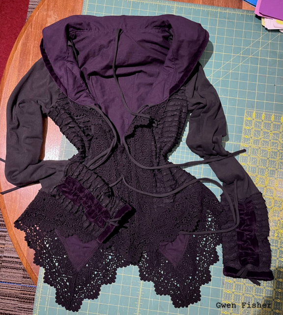 Photo of a black and dark purple hoodie jacket. It’s has a wide lace trim and a pointed hem. The front has straps to tie it shut. It’s gothic in style, suitable for a witch. 