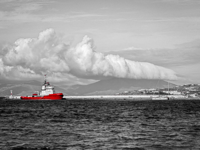 A black and white image of a bay with only a red ship to the left in colour and low hanging clouds as a backdrop. As seen from Little Venice, Mykonos