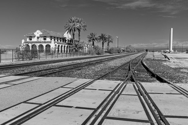 A black and white landscape photo of a old Spanish style train station in a desert landscape. The station is on the left. The view is looking down along four sets of railroad tracks. Two tracks join in an upside down shape just in front of the photographer. Five tall palm trees, a tree with a very long trunk and a tuft of long wide stiff green leaves at the top, line a fence that is between the station and the tracks on the right. 