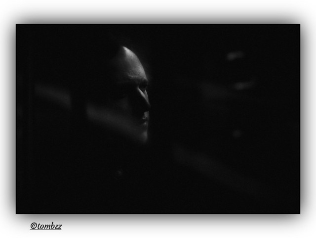 A black-and-white analog photo shows a man sitting by a window, wrapped in shadow. Soft light brushes the left side of his face, tracing his forehead, nose and cheek, while the right side fades into dimness. The shot is taken from outside, through the glass, with vertical and horizontal window bars framing the scene. His expression is quiet, maybe lost in thought, hard to read. The background disappears into darkness, adding to the feeling of isolation and tension. The whole image feels moody and still, like a moment caught between inside and outside, light and shadow.