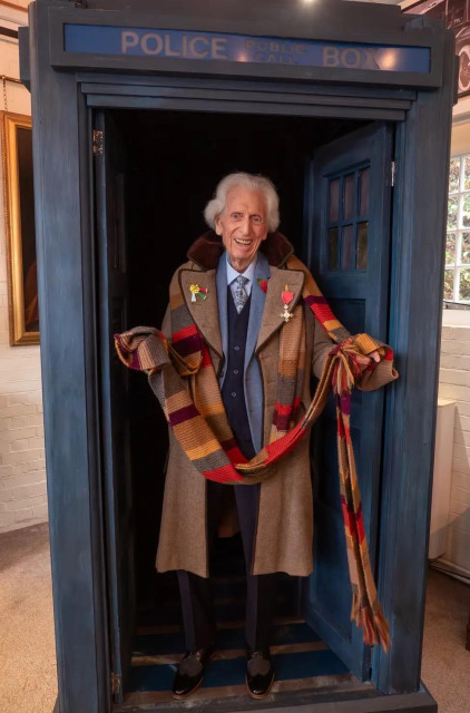 Tom Baker at 91 dressed as the Fourth Doctor holding The Scarf standing in the TARDIS doorway with his MBE pinned to his left lapel