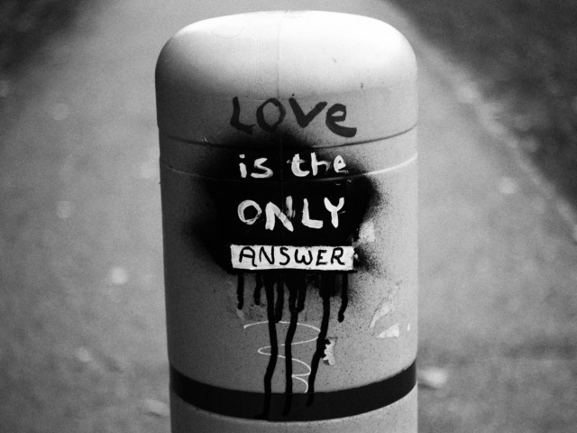 A protective barrier along a footpath is seen with graffiti on it saying "Love is the only answer". This is a black and white image.