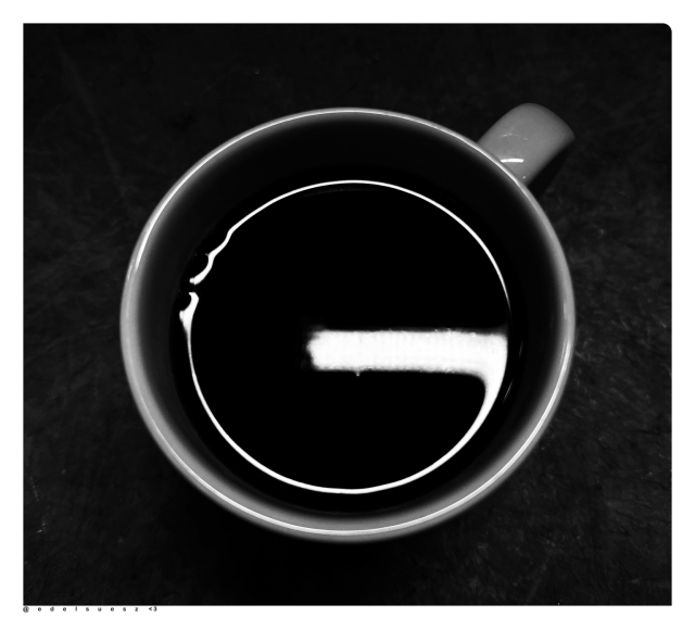 Aufnahme in schwarzweiß, Blick von oben in eine große runde gefüllte Kaffeetasse, die Umgebung ist dunkel, auf der Oberfläche des schwarzen Kaffees reflektiert sich eine G-förmige Lichtspiegelung 