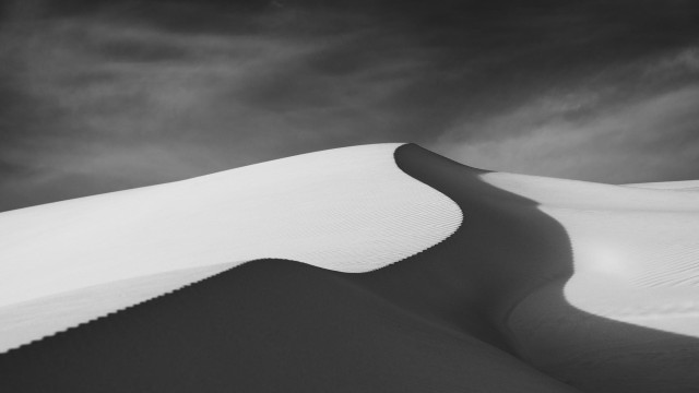 black and white dune photos with a sharp edge dune winding from bottom left via the center and into distance. The sky is dark with some light clouds.