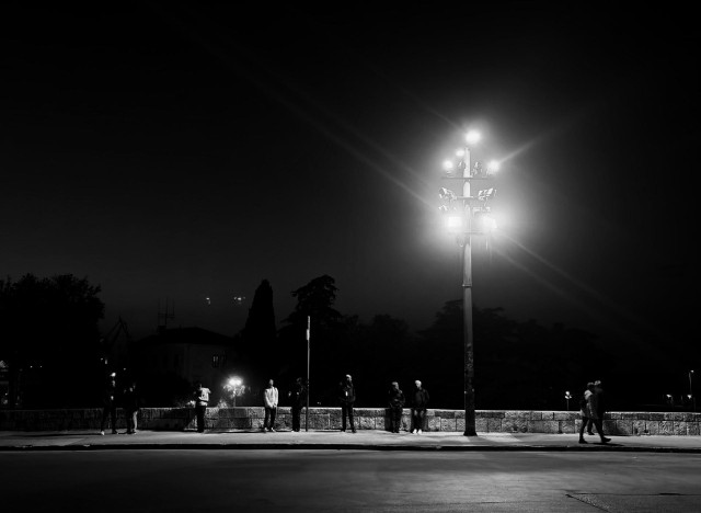 A nighttime photo looking across a street showing a row of people standing on a sidewalk along a stone wall illuminated by a tall streetlamp with multiple bright lights.