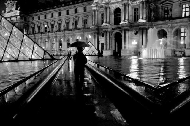 Une photo en noir et blanc au format paysage. Dans la cour du Louvre, de nuit mais éclairée. Au fond un des bâtiments anciens et sur la gauche l'avancée d'un angles de la pyramide en verre . L'avant plan de la photo, très sombre, montre une allée noire moirée de reflets dus à la pluie, elle court entre deux bassins dont l'eau lisse reflète les lumières du lieu . Au centre de l'image, se découpant contre l'une des petites répliques de la pyramide se détache la sombre silhouette  d'un visiteur sous son parapluie. 