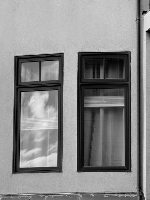Black and white study. Two narrow windows with smaller, two-pane windows on top. The trim is dark against a paler wall color. Both windows have those blinds you can raise from the bottom and lower from the top, each one bunched in the center of the window at different heights. Left side window reflects the sky with puffy clouds. Right side window is not reflecting, you can see through to the curtain hanging on the other side of the blinds. Nov 2025 SF