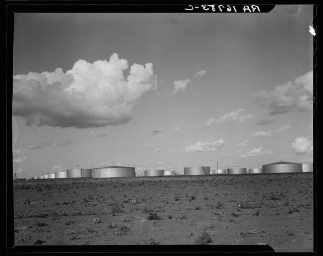 The black and white photograph depicts a series of large cylindrical storage tanks in an open field. These appear to be industrial oil or chemical storage facilities, with each tank having a domed top for containment purposes. The foreground shows sparse vegetation on dry ground that suggests arid conditions typical of the American Southwest. Above, there is a partly cloudy sky indicating variable weather. There are no visible people; only the tanks dominate the scene, conveying an industrial landscape. A handwritten code at the bottom right corner reads 'D-85231 RR,' which could be indicative of cataloging or archival information from the photographer's archive. The image captures a moment in time with significant historical and environmental context given its association with oil storage facilities during a period possibly related to World War II, as suggested by the code format that resembles wartime documentation systems used for photographs taken across various regions including Texas where Odessa is located.