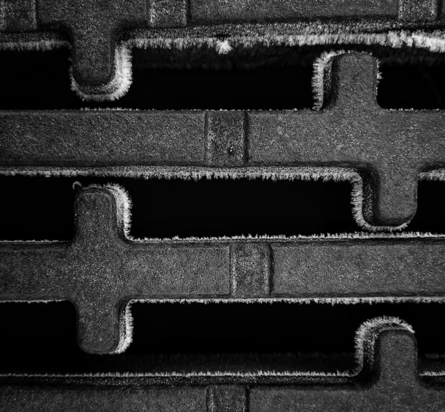 Close-up of a drain cover where the edges are covered in ice spikes. The picture is in b/w
