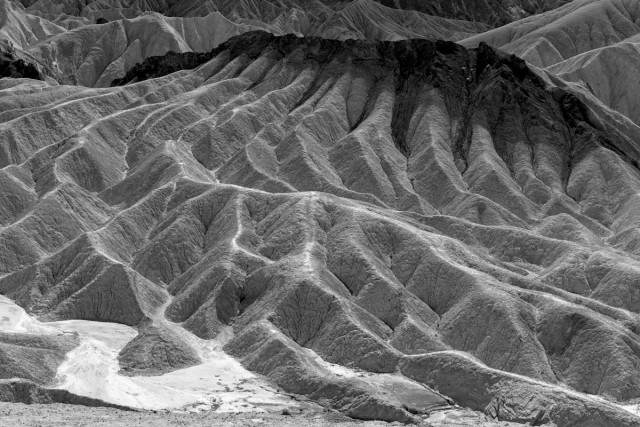 A black and white landscape photo of a barren desert hillside feature. The hill is completely barren and appears to be made of a fairly fine grain light colored material. The hill has many eroded valleys and crevasses. The top of the hill, however, is a very dark material adding contrast between the top and the rest of the hill. 