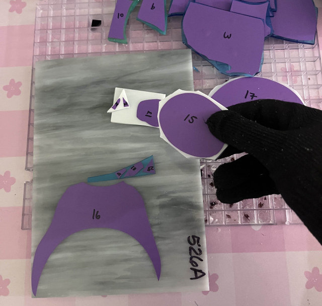 Right hand with cut proof gloves on holding a piece of white glass with purple vinyl labeled 15. In the background, there is a pile of broken glass and a grey glass with a template ready to be cut.