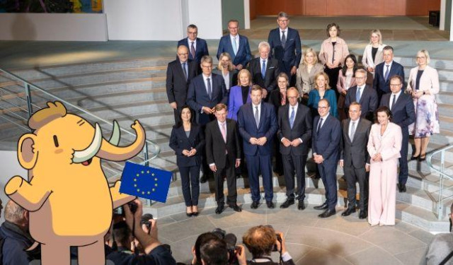 Das Bild zeigt ein Gruppenfoto der Bundesregierung, die in Richtung auf das in das Bild integrierte Mastodon-Maskottchen schauen. Es hält eine Europafahne in der Hand. 