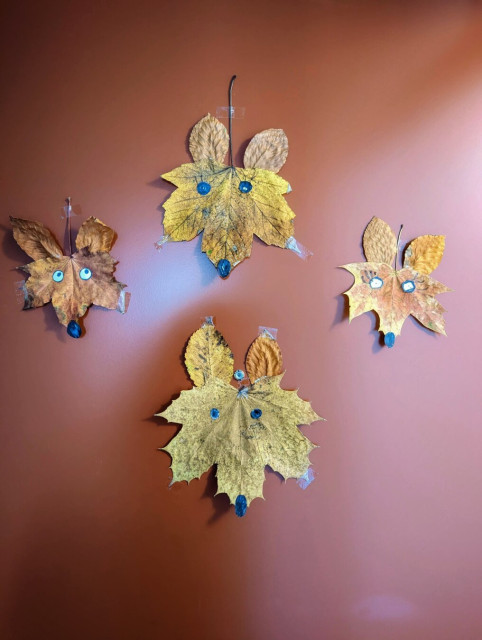 A playful door decoration featuring four leaf foxes crafted from dried autumn leaves. Each fox is assembled with a large leaf forming the body and smaller leaves as ears, showcasing a mix of golden-brown and warm orange tones. The foxes’ eyes and noses are either drawn on or made from small black and white paper cutouts, giving them a lively and endearing expression. The foxes are affixed to a reddish brown door.