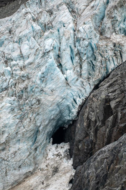 Close-up view of the Gabriel Icefall on the Gulkana Glacier, showing jagged blue-white ice fractured into steep seracs. A dark cave opens at the base where the glacier meets a vertical gray rock cliff. Streaks of dirt and old snow mark the lower ice, contrasting with the sharp texture and deep crevasses higher up the slope.