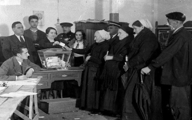 Three older Spanish women in long gowns voting at a polling station in 1933. There is a woman receiving their ballots, and a couple of men and a young woman watching.