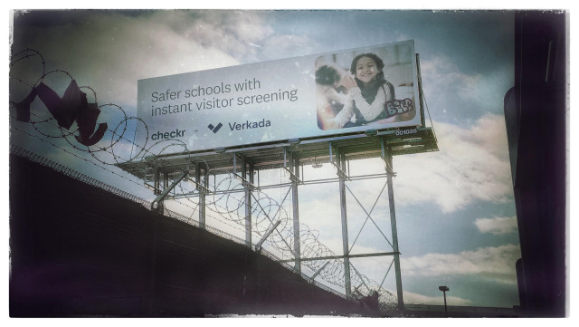 A view of a large advertising billboard, taken from street level next to a freeway in a grittier part of San Francisco. 

The billboard promises "Safer schools with instant visitor screening" ... and it has a picture of a smiling child. The picture has been tagged with graffiti... and underneath the billboard there's a fence topped with barbed wire.

End Times are approaching. I have my camera ready 👍
