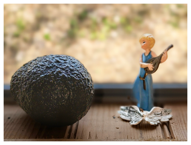 	
A rough-textured avocado sits on a wooden surface next to a small ceramic figurine of an angel in a blue dress, holding a guitar with their detached wings lying in front of them. The background is softly blurred, showing light colors and some greenery.