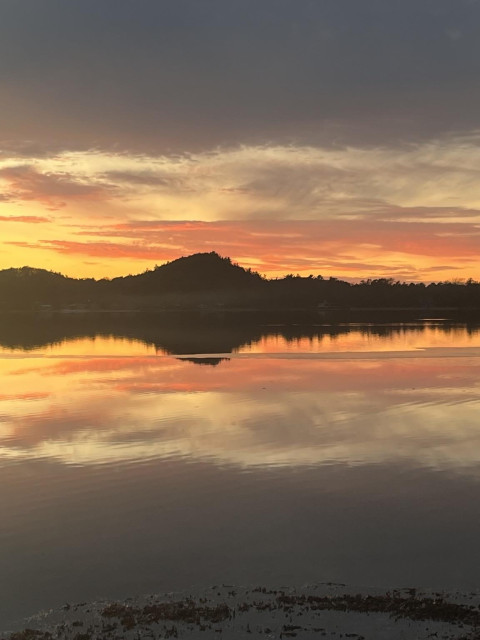 A pastel sunset of orange, yellow and red is reflected on a calm lake with dark sand dunes on the horizon, creating a symmetrical picture. Dark grey clouds are along the top and reflected on the bottom, giving the illusion that the strip of color in the middle of the picture is a torn piece of paper.