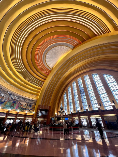 The image shows the interior of the Cincinnati Union Terminal's main concourse. This iconic building, an Art Deco masterpiece, is a National Historic Landmark in Cincinnati, Ohio. 
Opened in 1933, the terminal is famous for its massive half-dome ceiling, which was the largest in the Western Hemisphere at the time. The interior features curving, layered bands of stucco painted in yellow, orange, and silver tones, characteristic of the Art Deco style. 