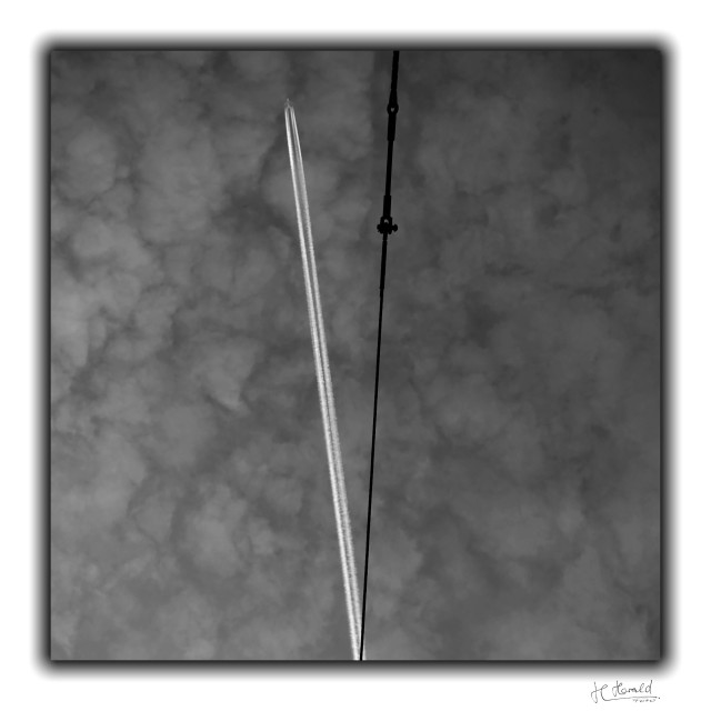 Square black-and-white image of a cloudy sky. Thin clouds cover the entire sky and look a bit like fleecy clouds. In the centre at the bottom, a white quadruple contrail extends slightly to the left and a thick black tension cable extends slightly to the right from the same point. Both form a ‘V’ shape.