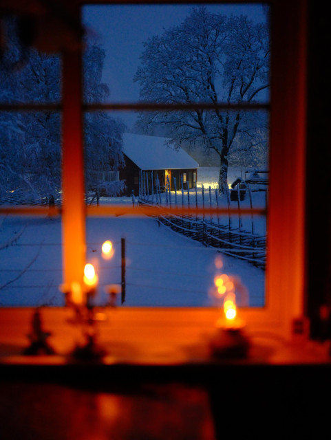 Winter landscape of a rural farm in Sweden in blue hour. A snow covered barn seen through a window lit by candles.