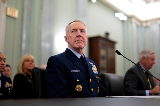 Older clean-cut white man in a decorated military uniform, sitting at a table with a microphone pointed at him in a confirmation hearing. His facial expression tells the story of a smug nazi.