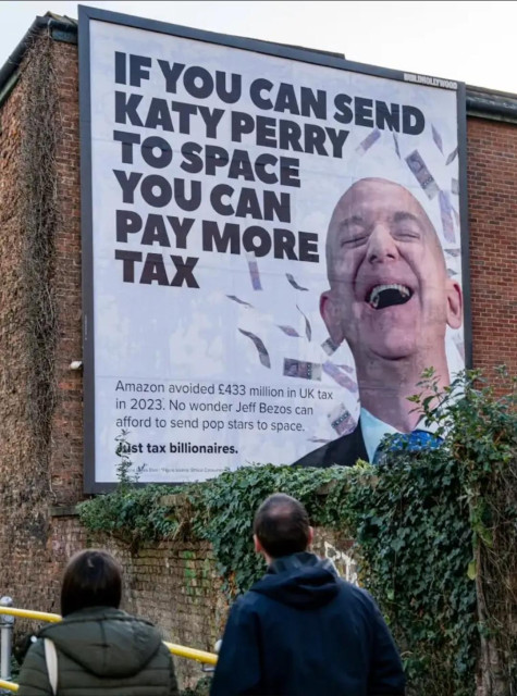 A photograph shows a large billboard with a portrait of a man in a suit. The man is smiling and appears to be looking slightly upwards. Scattered around the man’s head are images of dollar bills. Below the portrait is a white rectangle with black text that reads “Amazon avoided £433 million in UK tax in 2023. No wonder Jeff Bezos can afford to send pop stars to space. Just tax billionaires.” In the foreground, two people are standing and looking at the billboard. The billboard is mounted on a dark gray surface, and there is a metal railing in front of it.

Provided by @altbot