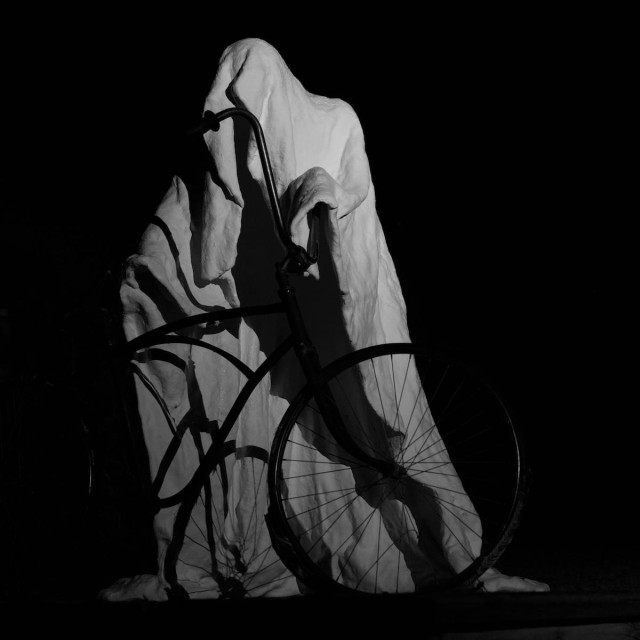 A square black and white photo of a sculpture at night. The sculpture appears as a person in a long white flowing cloak. Think classical monk with the a long hood that covers the face. The ghostly figure is holding on to a bicycle as if it were going to mount the bike and ride off. The sculpture is fairly well lit but the bike is shadowy. Otherwise, the rest of the frame is black.