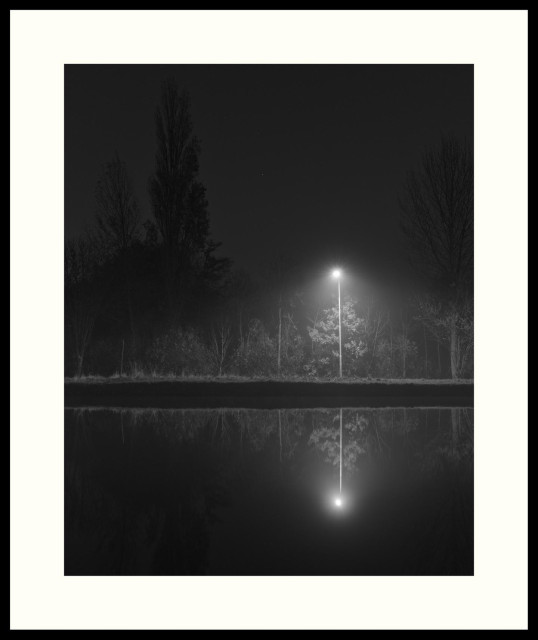 b&w - A single street lamp illuminates part of the opposite side of a canal. The grass, bushes and lower parts of trees are visible in the spread out cone of light. Outside and behind the light silhouettes of tall trees can be seen. A few stars are still visible in the sky. The complete absence of wind creates an almost perfect reflecting  mirror. Shot on a very early and somewhat hazy morning.