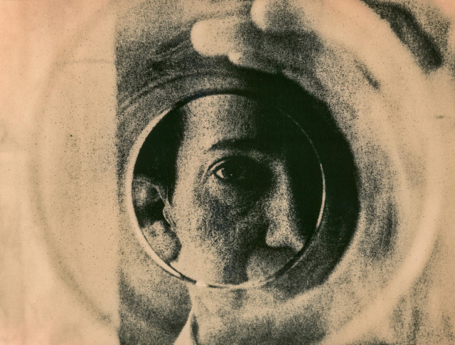 This lith print depicts a portrait seen through a drinking glass held up by the person themselves. The circular rim of the glass frames the face, distorting it slightly and creating ripples of soft, swirling tones around the central eye. The heavy grain and high contrast typical of lith printing intensify the sense of texture, giving the image a tactile, almost atmospheric quality.

The visible eye, emerging sharply from the darkened interior of the glass, becomes the focal point—quiet, direct, and introspective. The surrounding shapes blur into abstract rings, as if the world outside the glass dissolves into soft haze. The result is a portrait that feels both intimate and experimental, blending human presence with analogue distortion to evoke a sense of self-observation and quiet mystery