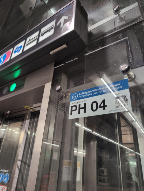 Außenansicht eines Bahnsteiglifts der Wiener Linien. Wände, Türen und Liftkabine sind aus Glas; Ränder, Rahmen udgl aus grauem Metall. An der Wand neben der Tür ist ein neues Hinweisschild zu sehen, auf dem sehr groß "PH 04" steht (das ist die Bezeichnung dieses Aufzugs), dazu ein QR-Code. Darüber steht kleiner "Aufzug barrierefrei bedienen / Accessible control for elevators. Unter dem QR-Code steht klein sein Inhalt: wienerlinien.at/Liftboy