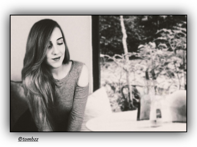 A young woman sits at a round table, facing the light. Her eyes are closed, hands likely folded. A gesture that hints at focus or quiet. There's a faint smile on her lips, subtle and uncertain, somewhere between contentment and deep thought. She's wearing a light top with open shoulders and a short skirt, and she seems wrapped in a kind of inner calm. Behind her is a white wall, and further back, a large window looking out onto a garden. Leaves, branches, soft light. It's an analog black and white portrait, gentle in tone and quiet in mood. The woman remains a mystery.