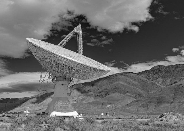 A black and white landscape photo of a very large radio telescope dish. The dish sits on a single pedestal. It points up and toward the right. Sagebrush is in the foreground and a large mountain in the background. Big clouds in the sky frame the outline of the telescope's four pyramidal feed support legs. The cloudless part of the sky is very dark as if a red filter had been used.