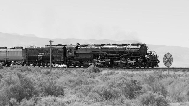 A black and white landscape photo of a very large black steam engine in a desert landscape. The engine is heading toward the right side of the frame. It is very long, with two sets of drive wheels. The tender car is behind the engine and a small section of a passenger car can be seen behind the tender.  Sagebrush is in the foreground, and dimly, through the haze, a mountain range is seen in the distance. A round, railroad crossing sign is seen just ahead of the engine on the right. It is filled with bullet holes. 