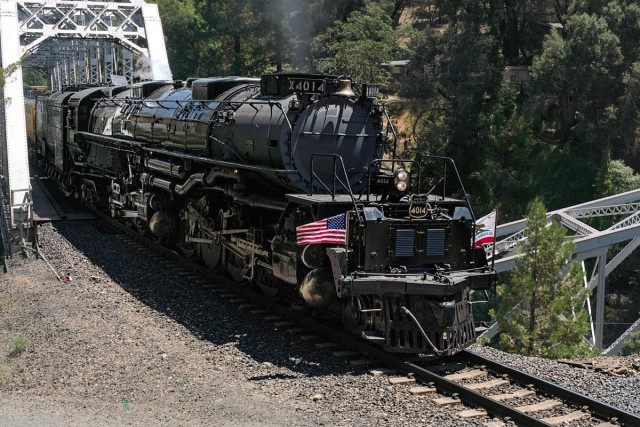 A color landscape photo of a very large steam engine. The engine is stationary just as it cleared a bridge. The rest of the train is still on the bridge. Another bridge can be seen crossing under the upper bridge where the tr5ain is on the left. The train points toward the right. Whisps of black smoke come from the front smoke stack. It has a small US flag on the right front and a California flag on the left front. A forest is seen in the background.