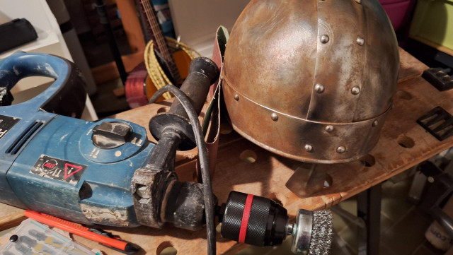 A rusty, norman-style helmet, quite rusty, and a electric drill with metal brush attached. 