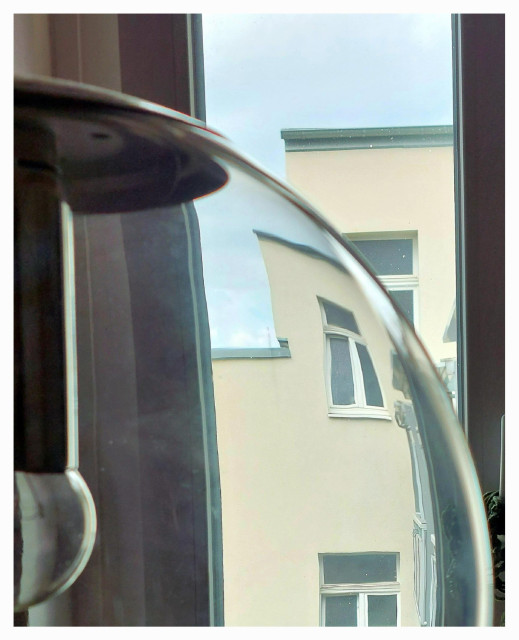 View through a glass lamp from a room window onto a house opposite. The lamp, of which only the right half is visible, is a spherical ceiling lamp made of colourless glass, with a silvery bulb and dark socket. Due to the curvature of the glass, the house façade with its windows appears convexly curved and bent. Above the house is a bluish sky.