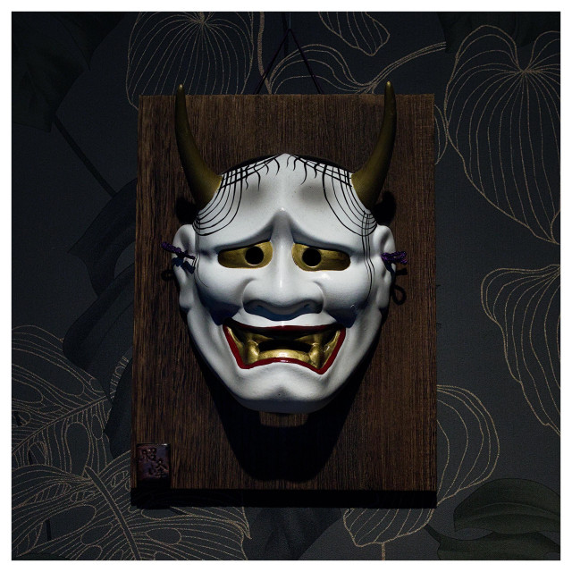 En fyrkantig bild på en vit mask med horn. Masken har röda läppar och stora tänder och ser skräckinjagande ut och sitter på en träbit, upphängd på en vägg med mörk, mönstrad tapet.