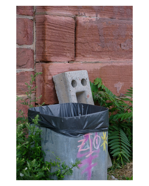 Foto im Hochformat. Ein öffentlicher Mülleimer vor einer roten Backsteinwand. Links und rechts davon sind grüne Pflanzen. Schräg hinter dem Mülleimer steht ein Betonklotz. Dieser hat oben zwei Löcher nebeneinander. Darunter ist eine rechteckige Öffnung. Es sieht aus wie ein Gesicht mit aufgerissenem Mund.