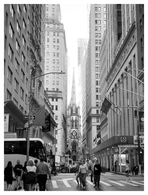 Black and white photo of Trinity Church in NYC seen at a distance through a canyon of tall buildings, with pedestrians and street traffic in the foreground. The church steeple appears to be squeezed in by buildings on both sides, and the windows and a clock on its facade resemble a narrow face with an open-mouthed shocked expression.